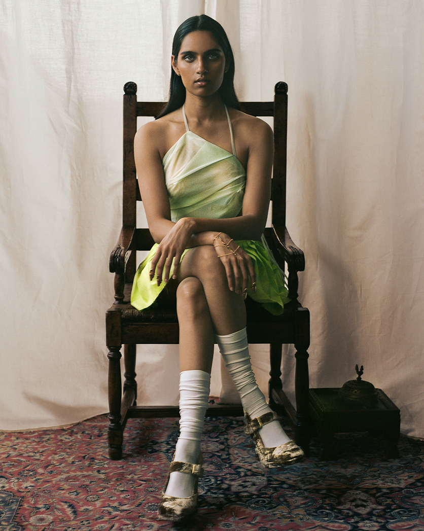 Model sitting on a wood chair in a green top and skirt looking directly at the camera with smokey eye makeup